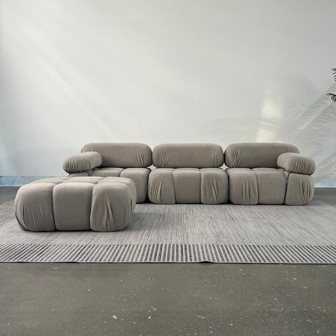 A beige modular sofa system with a plush appearance, consisting of multiple cushioned units and an ottoman, arranged on a grey rug.