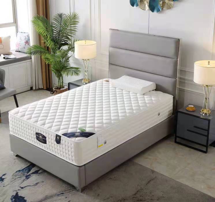 A white, patterned mattress on a gray bed frame, placed in a bedroom setting with decorative pillows, a nightstand, and a lamp.